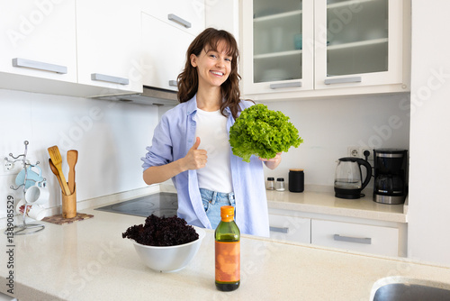 Beautiful curly brunette holding green salad, smiling and showing thumbs up in the kitchen at home