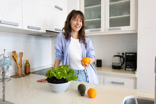 Curly smiling brunette winking and holding an orange and getting ready to cook a dinner with fruit and veggies in the kitchen at home