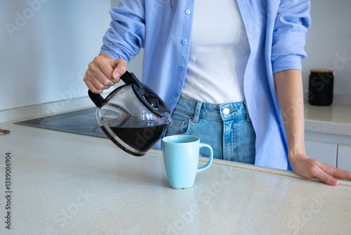 Woman holding a coffeepot and pouring coffee into a cup in the kitchen at home in the morning