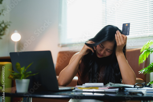 Young stressed asian woman calculating monthly home expenses holding credit card having financial problems and using laptop working from home