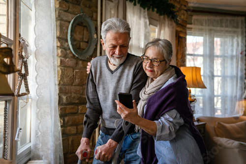 Obraz na płótnie Senior wife embracing disabled husband at nursing home. Cheerful old couple using mobile phone. w salonie