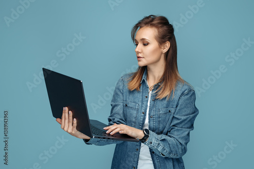Serious young woman in jeans shirt using computer or laptop over blue background