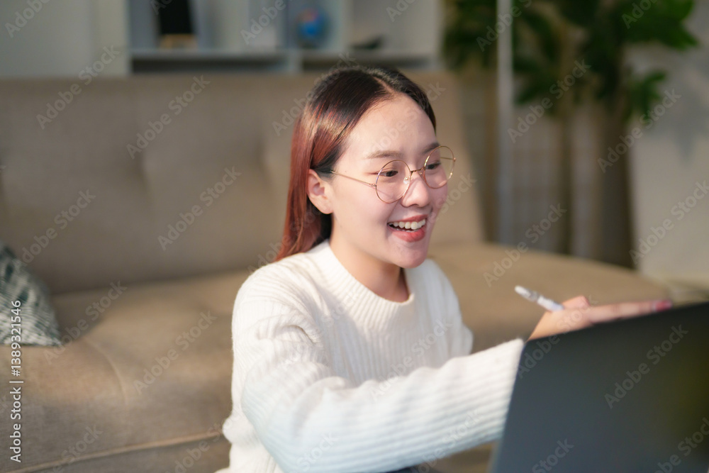 Obraz premium Smiling Asian freelancer working remotely from home, using a laptop in a cozy living room, balancing productivity and comfort