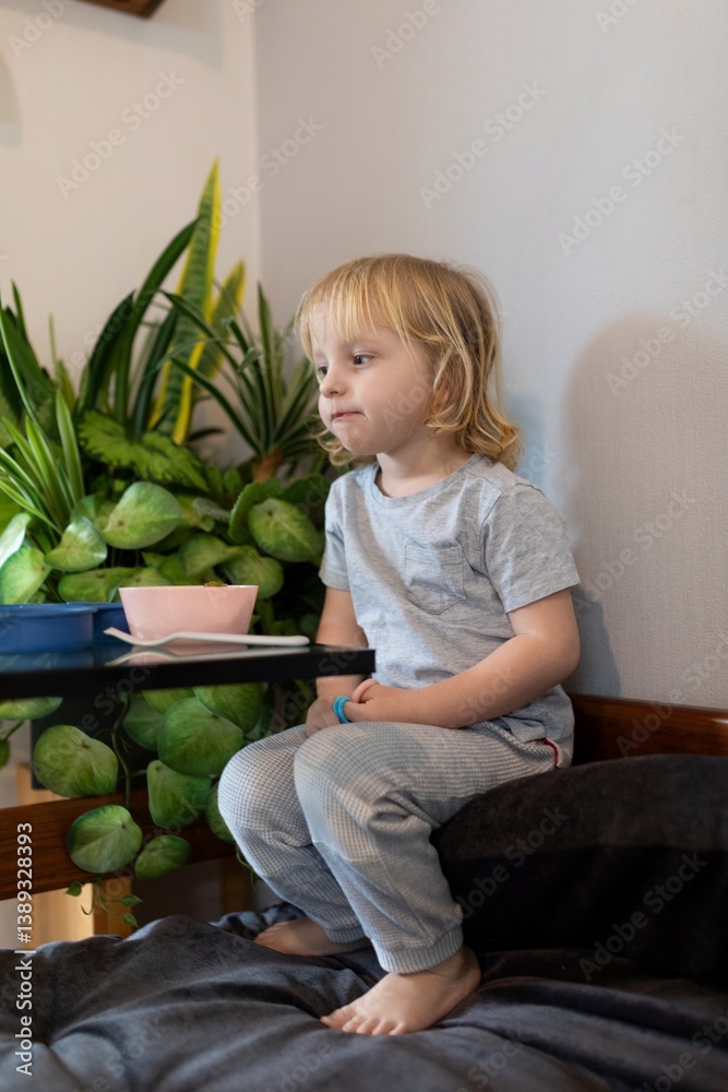 Young child sitting quietly on a couch next to a green potted plant in a cozy indoor space during the day