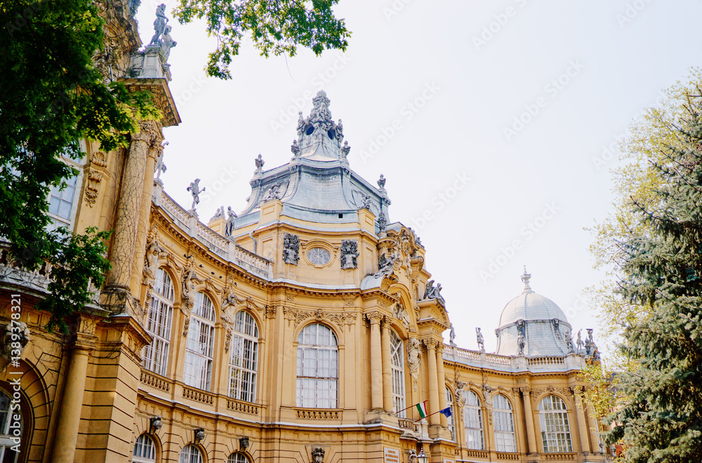 Fototapeta premium A beautiful historic building with ornate architecture, featuring intricate sculptures and a domed roof. Surrounded by lush greenery, the structure showcases large windows and decorative details.