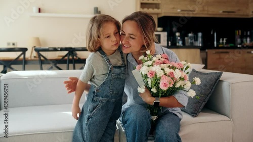 Young boy gives his mother a greeting card and flowers at home in a cozy setting. She reacts with joy and warmth. Concept of Mother's Day celebration and everyday family love. Mid-range shot natural