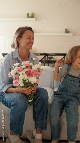 Young boy gives his mother a bouquet of flowers at home in a warm and cozy setting. She smiles with joy and gratitude. Concept of Mother's Day celebration and family love. Mid-range shot natural light