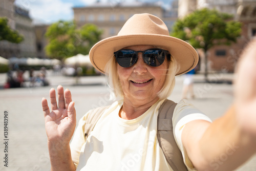 Obraz na płótnie Senior woman smiles while taking a selfie in the city square, waving her hand. She is wearing a beige hat and sunglasses, enjoying a sunny day out while sightseeing. w salonie