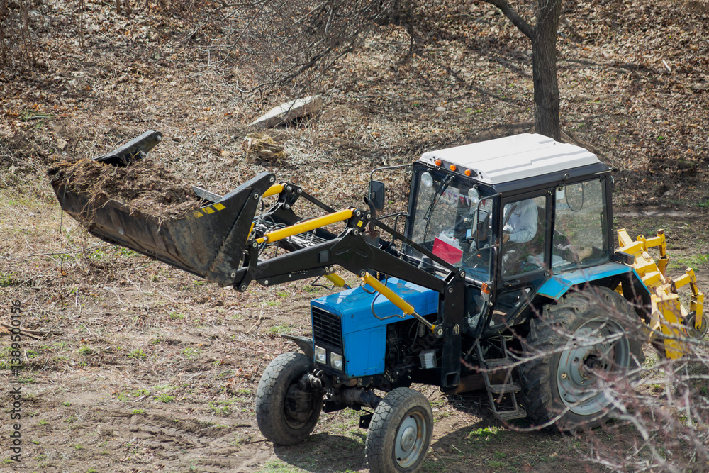 Obraz premium The construction loader with an elevated bucket, carrying debris and wood waste, is clearing the area. Landscaping work is in progress.
