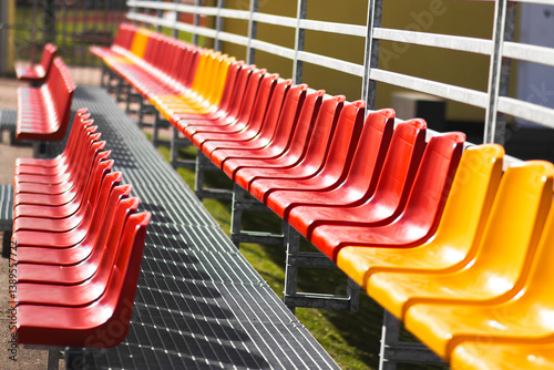 Red and yellow stadium bleachers on a sunny day. Empty spectator seats arranged in rows, creating a vivid geometric pattern. Outdoor sports arena background.

