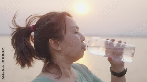 A close-up shows an overweight Asian woman in sportswear drinking water by the lakeside after exercising during sunset, a beginner caring for her health on her fitness journey.