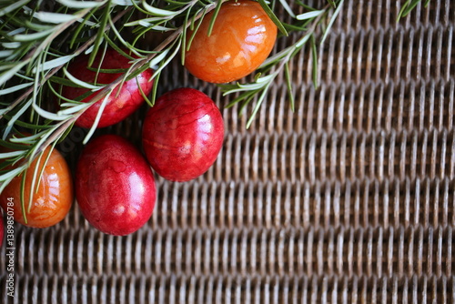 Easter eggs and a twig of rosemary are lying on the woven table. The light is beautifully reflected in the eggs through the balcony railing. Red and orange eggs.