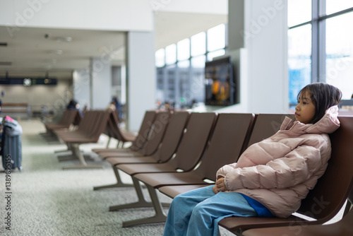 Asian adorable kid girl traveler in winter jacket sits alone in airport lounge, lost in thought. The stillness, vacant chairs, and soft lighting capture solitude, patience, and the journey ahead.