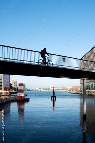 Cyclist riding over water on bridge in Copenhagen, Denmark, promoting green commuting and smart urban design aligned with environmental sustainability and innovation.