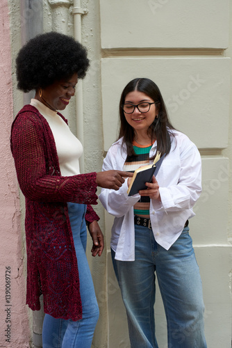 An African-American woman and an Asian-American woman smiling and discussing over a notebook on a city street corner