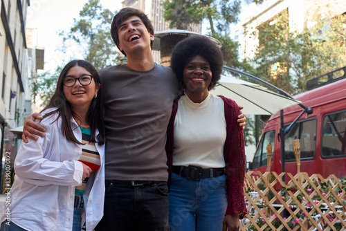 Three friends standing together, smiling and laughing, in a lively city street with outdoor seating