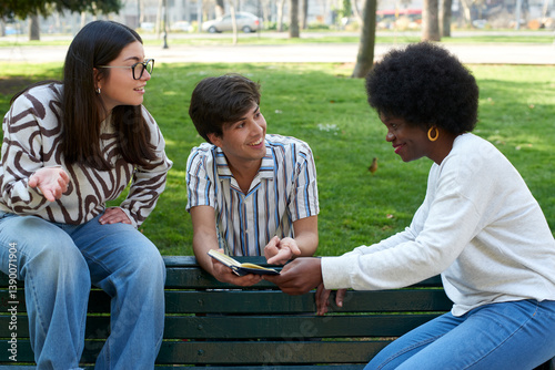 Three friends sitting on a park bench, smiling and interacting while looking at a paper notebook
