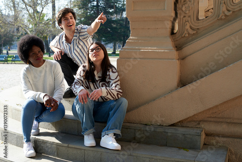 Three friends sitting on steps outdoors, with one pointing in the distance while enjoying a casual moment together