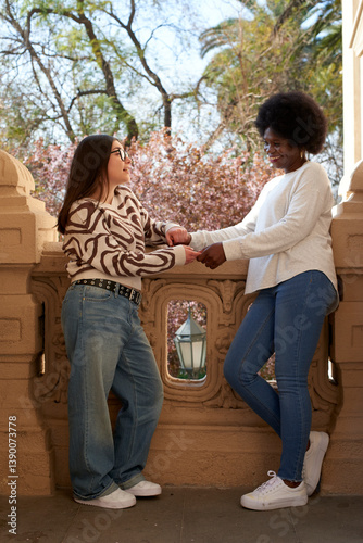 Two women standing on a terrace, holding hands and smiling, with trees in the background