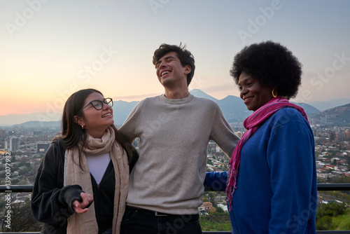 A group of friends smile and embrace while enjoying the sunset over a city, creating a joyful and warm scene