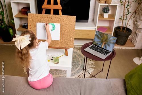 Girl recreating palm tree from laptop screen, using green paint, studying nature through digital art tutorial in relaxed home setting. Concept of childhood, online education, remote studying