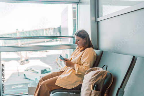 Obraz na płótnie A young traveler in casual yellow attire sitting in the airport lounge using two mobile phones while waiting for her flight. Perfect for topics related to modern travel, technology, and leisure time. w salonie