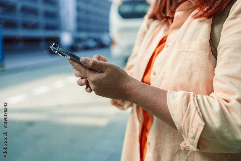 Woman with red hair is holding her smartphone in hand while looking at the screen outdoors. This image highlights the use of smartphones for digital interaction, messaging, or navigation in urban
