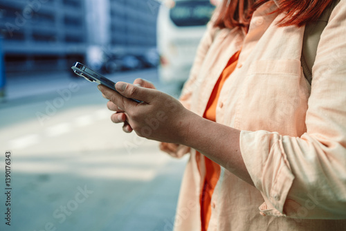 Woman with red hair is holding her smartphone in hand while looking at the screen outdoors. This image highlights the use of smartphones for digital interaction, messaging, or navigation in urban