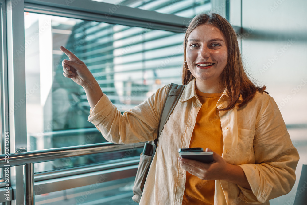 A cheerful young woman in a yellow shirt and jacket standing at the airport terminal. She is pointing at something with a smile, holding a smartphone in her hand. Ideal for travel, tourism, and