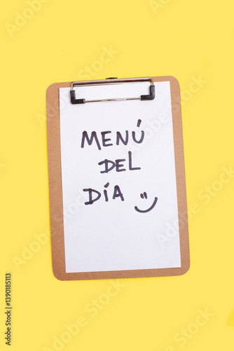 Menu del dia and a smiley face written on a white sheet on a clipboard on a yellow background (trad. menu of the day)