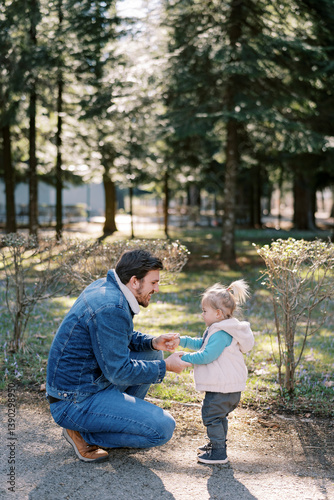 Dad holding hands a little girl squatting next to her in a spring park