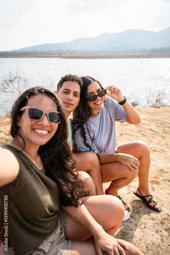 Latin hispanic friends taking a selfie by a scenic lake surrounded by nature and mountains at summer