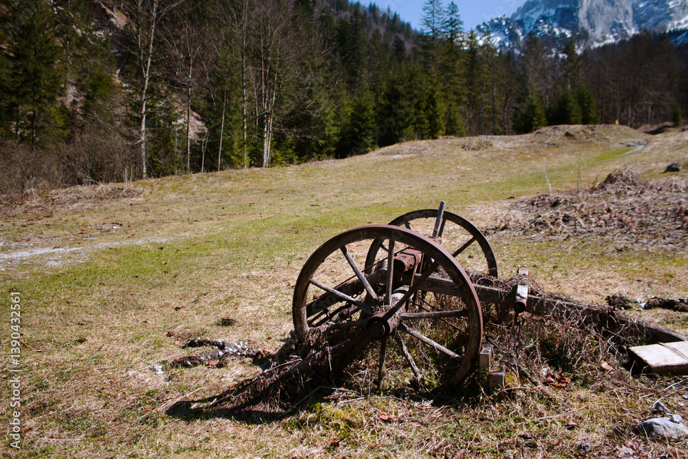 Fototapeta premium Old wagon wheel rests in a mountain valley with snowy peaks.