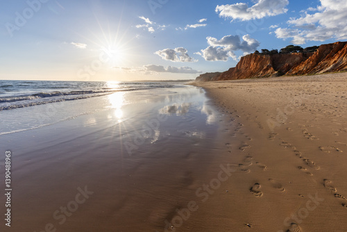 The golden sand beach of Praia da Falésia, crystal-clear sea water at low tide with the reflection of the sky, the setting sun, and the characteristic orange cliffs, Algarve, Portugal