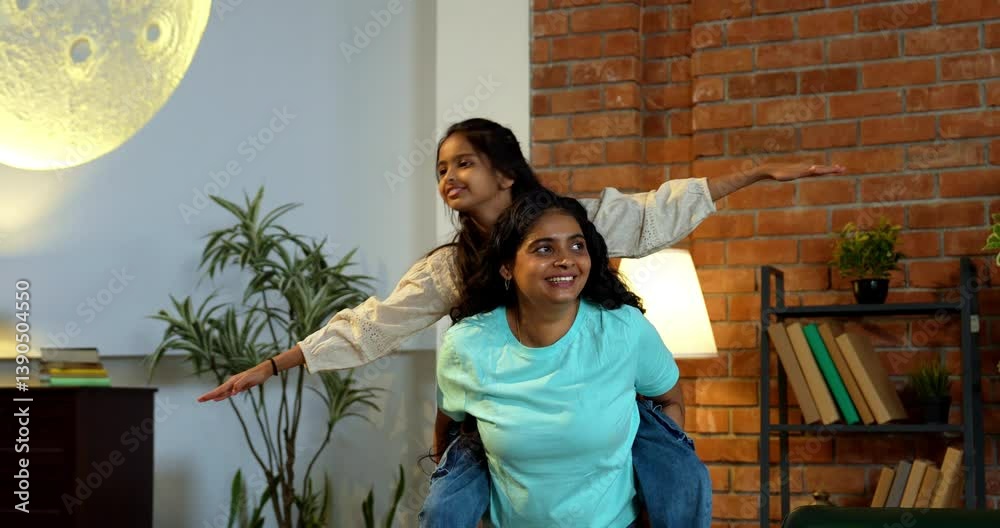 Indian Asian cute little girl joyfully riding piggyback on her mother’s back in a modern living room, both laughing and enjoying playful quality time together, creating a loving, happy family moment