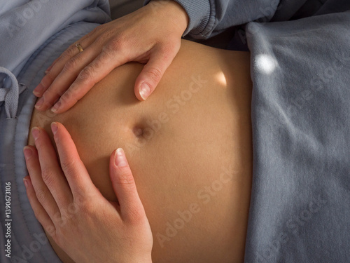 Pregnant woman rests calmly on bed, gently touching belly with loving hands. Pregnant moments filled with peace and hope reflect beauty of being pregnant and nurturing life inside. 