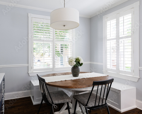 A cozy dining room with blue walls and white plantation shutters, black chairs and white bench seating around a wood table, and a white light fixture hanging above.