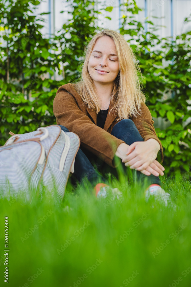 Young woman sitting on a grassy lawn with a backpack, brown jacket, black pants, background is garden or park with bushes. the overall mood of the image is peaceful and relaxed.