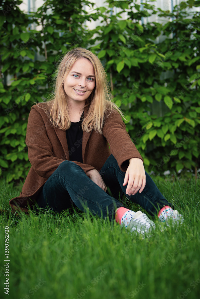 Young woman sitting on a grassy lawn with a backpack, brown jacket, black pants, background is garden or park with bushes. the overall mood of the image is peaceful and relaxed.