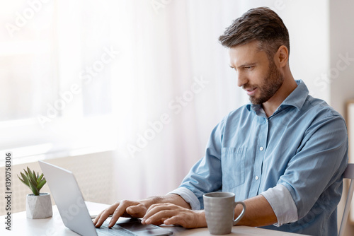 Unemployed man scrolling job search website at home using laptop computer, handsome male freelancer working remotely at desk near window, typing on keyboard, answering emails, copy space