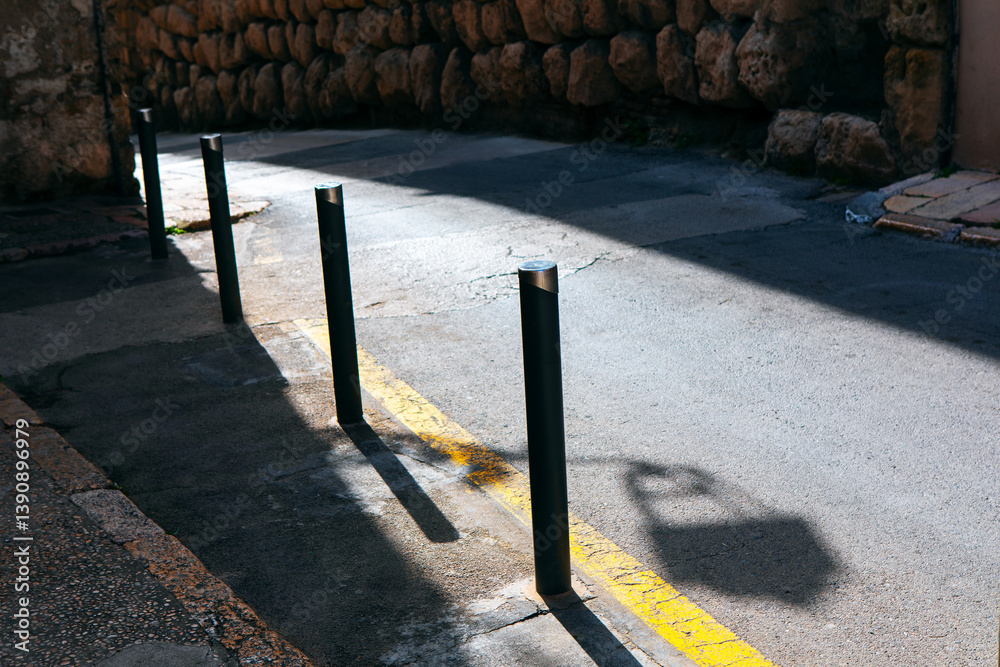 Narrow street illuminated by sunlight, highlighting bollards lined up along road marking. On one side, textured stone wall contrasts with smooth pavement, while shadows add depth to urban composition