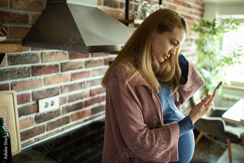 Obraz na płótnie Smiling pregnant woman using smartphone at home in modern kitchen w salonie