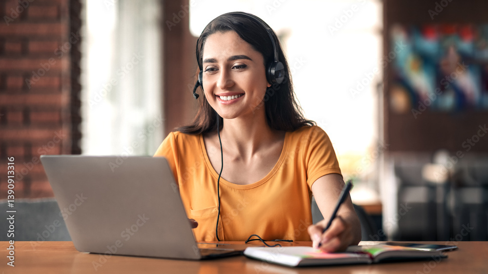 Fototapeta premium Happy young arab woman having educational course online, using laptop and headset, cafe interior, free space. Smiling female student studying online, sitting in front of laptop at cafeteria