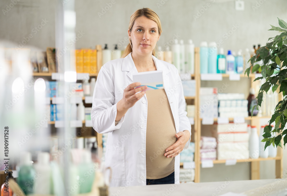 Friendly pregnant apothecary holding a box with remedy standing at the desk in drugstore
