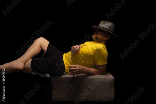 Mature man, 50s, quite uninhibited, with a beard and sunglasses, wearing a hat and yellow shirt posing calmly for a photo sitting on a beanbag.