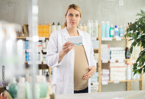 Friendly pregnant apothecary holding a box with remedy standing at the desk in drugstore