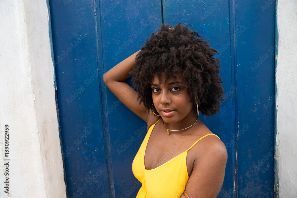Fototapeta premium Close-up portrait of a young woman with afro hair, wearing a yellow dress, sitting by a blue door of an old house, looking at the camera. Confident person.