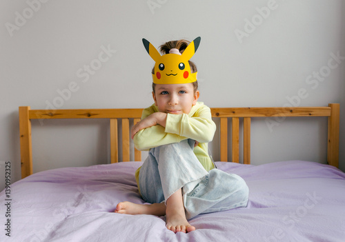 Cute little girl in a yellow hoodie and a Pokémon mask on her head is sitting on the bed.