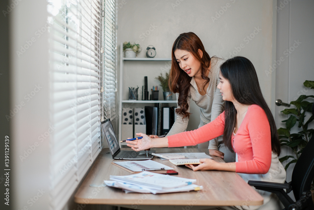 Two women working together at a desk, discussing documents and using a laptop in a bright, modern office environment. Two women working together at a desk, discussing documents and using a laptop in a bright, modern office environment.