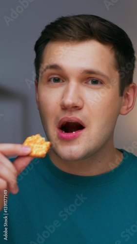 Portrait of Young Man Eating Sweet Food and Looking at Camera Close Up. Modern Person Bites Sesame Cookie and Relaxes in Joy. Appetite of Alone Hunger Guy with Crispy Treat in Living Room Interior